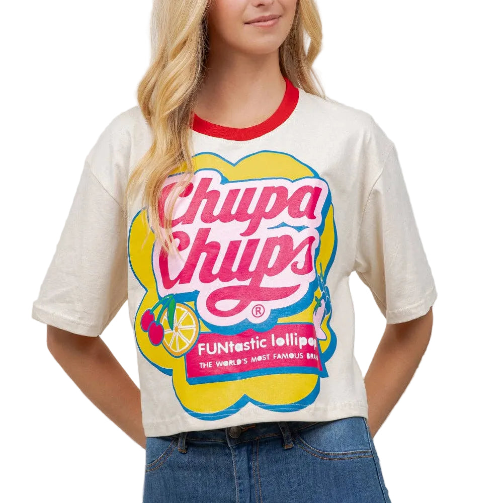 A woman wearing a cream-colored Chupa Chups graphic t-shirt with a red crew neck collar and blue jeans, standing against a light beige wall with a green plant on the right.