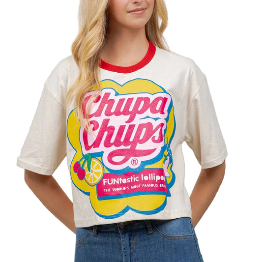 A woman wearing a cream-colored Chupa Chups graphic t-shirt with a red crew neck collar and blue jeans, standing against a light beige wall with a green plant on the right.