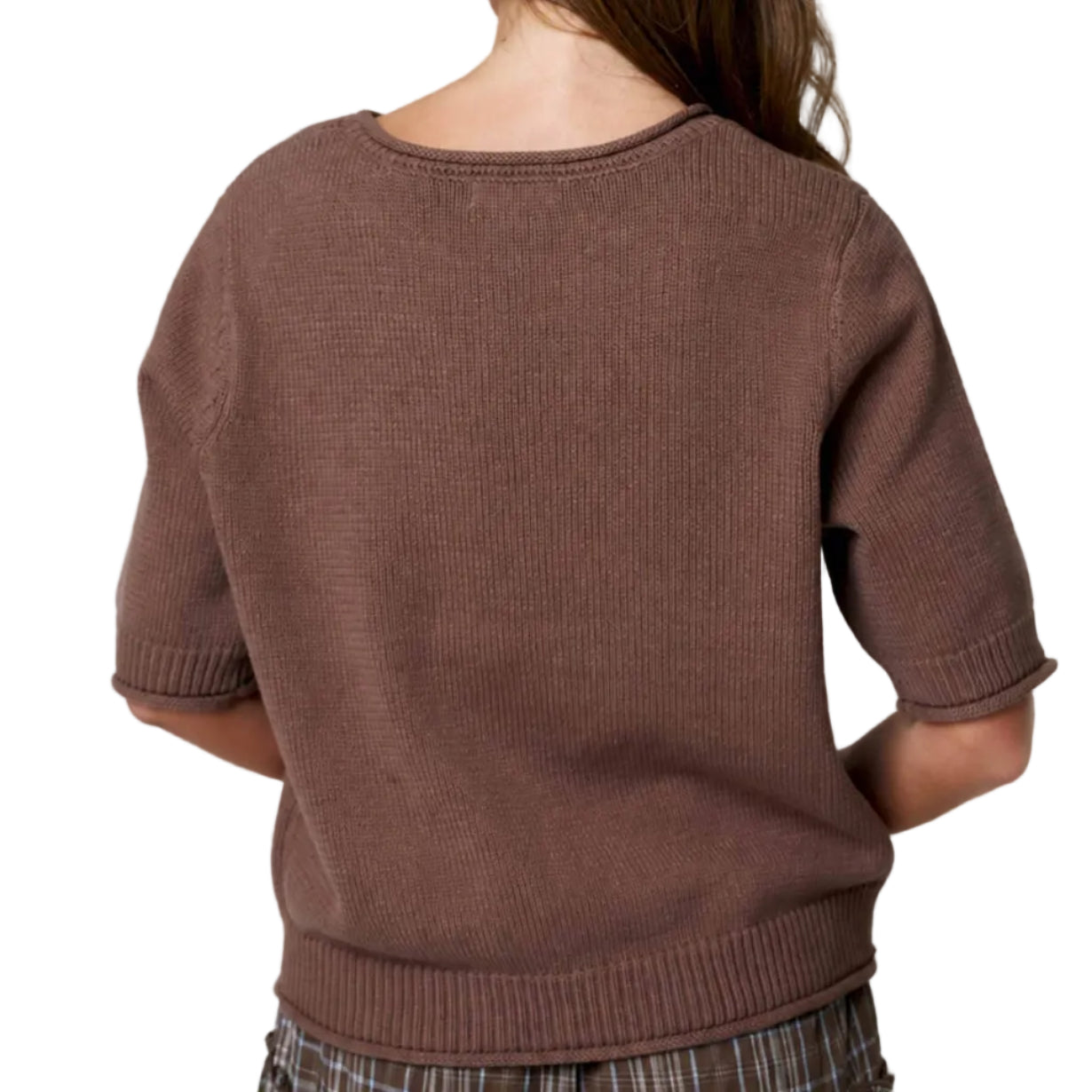 Person wearing a brown sweater and plaid skirt standing against a white curtain.