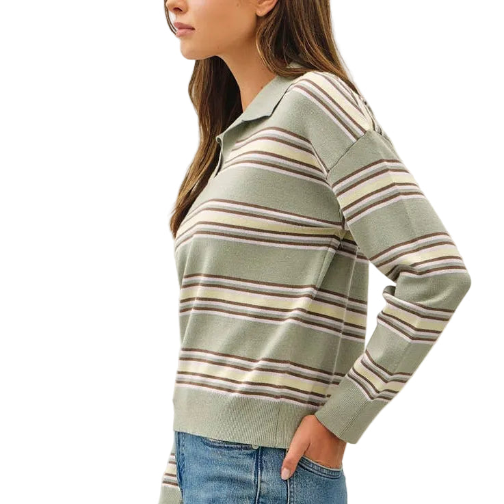 A woman modeling a sage green long-sleeve knit sweater with thin brown and cream stripes, paired with blue jeans and brown sneakers, standing against a plain off-white studio background.