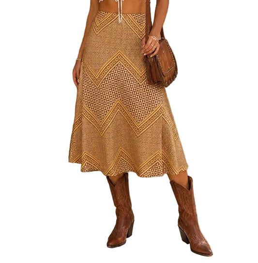 A woman stands outdoors wearing a bohemian-style outfit consisting of a white lace crop top, a tan and brown patterned crochet midi skirt, a beige wide-brim hat, brown cowboy boots, and a brown shoulder bag.