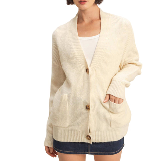 Woman wearing a cream cardigan over a white top and denim shorts on a plain background