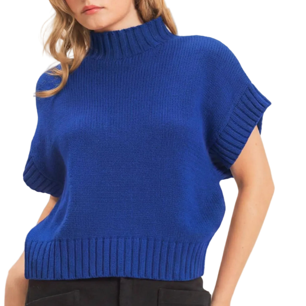 A woman wearing a vibrant royal blue ribbed knit mock-neck sweater with short dolman sleeves and black trousers, photographed against a light beige studio background.