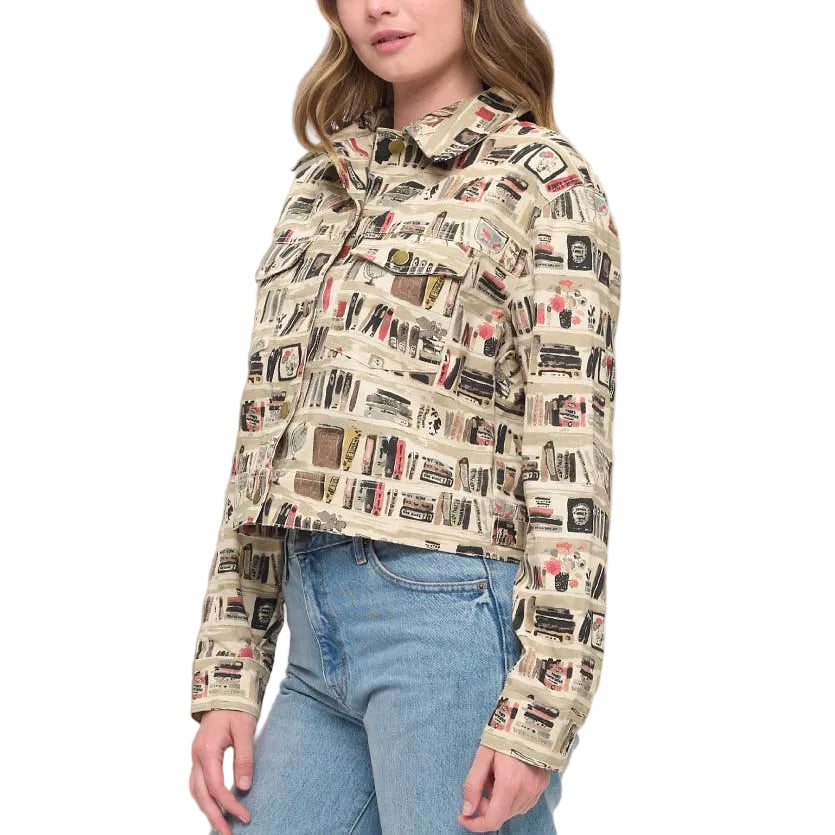 A woman wearing a beige long-sleeve cropped jacket with a detailed vintage book and illustration print, paired with light blue jeans, standing in a brightly lit room with a neutral beige wall and a decorative vase.