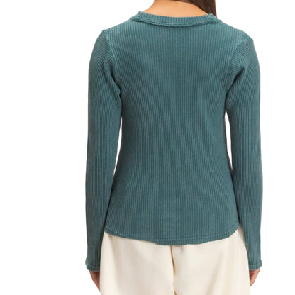 A woman viewed from the back wearing a teal ribbed long-sleeve top and cream pants against a plain beige studio background.