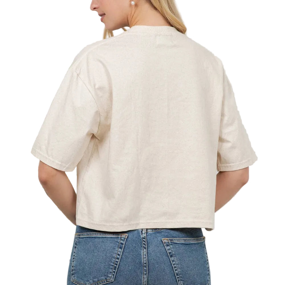 Woman wearing a beige top and blue jeans against a white wall with plants.
