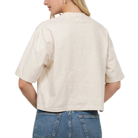 Woman wearing a beige top and blue jeans against a white wall with plants.