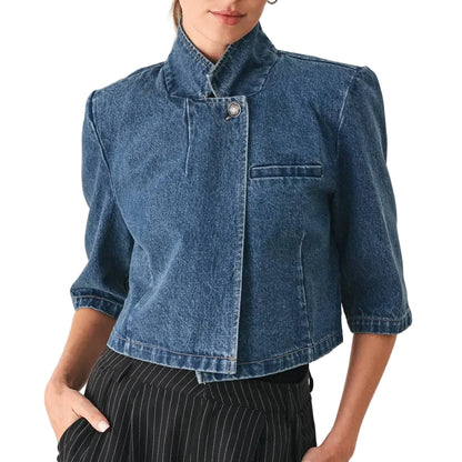 A woman modeling a cropped blue denim jacket with a high stand collar, structured shoulders, and elbow-length sleeves, paired with black pinstriped trousers against a light gray studio background.