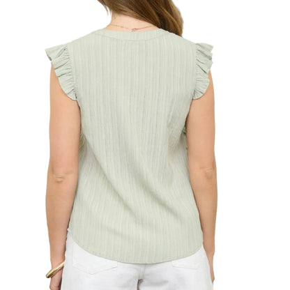 Woman wearing a light green sleeveless top with ruffled shoulders, standing in front of a white wall with plants.