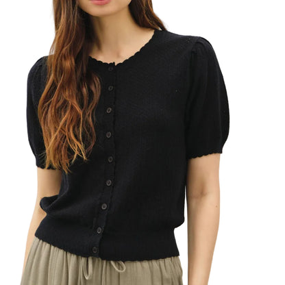 Woman wearing a black short-sleeve cardigan over a black top with beige pants on a white background