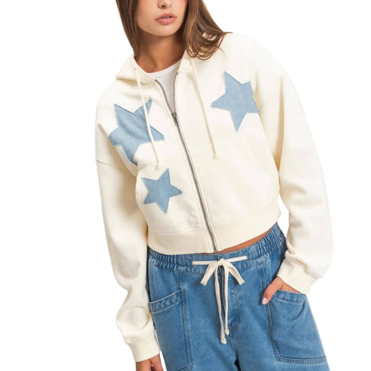 A woman wearing a cream-colored cropped zip-up hoodie with light blue star patches, paired with blue jeans, against a plain beige background.