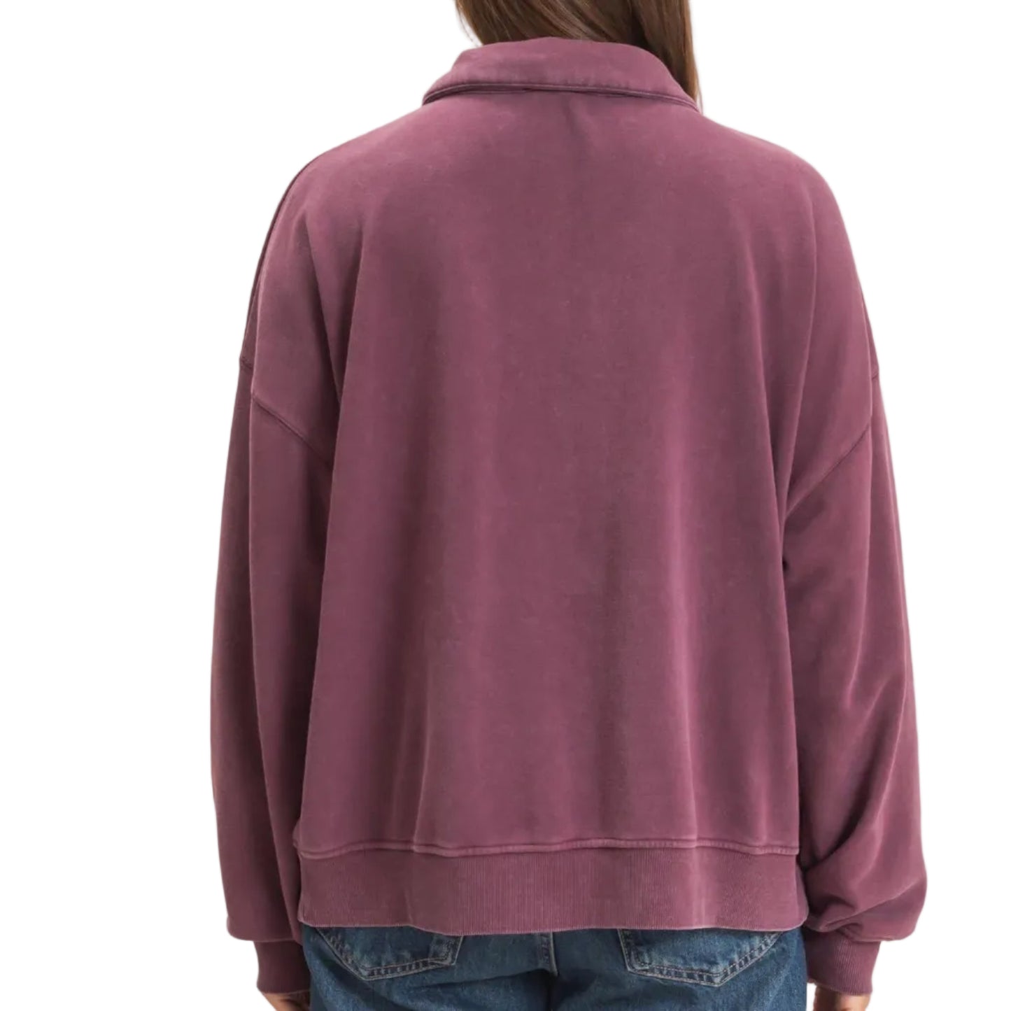A woman viewed from the back wearing a dusty mauve-purple fleece jacket and blue jeans, set against a plain beige studio background.