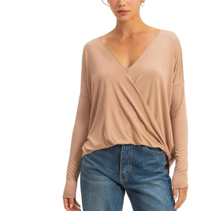 A woman modeling a beige long-sleeve wrap-style top with blue jeans and tan shoes, standing against a plain light beige studio background.