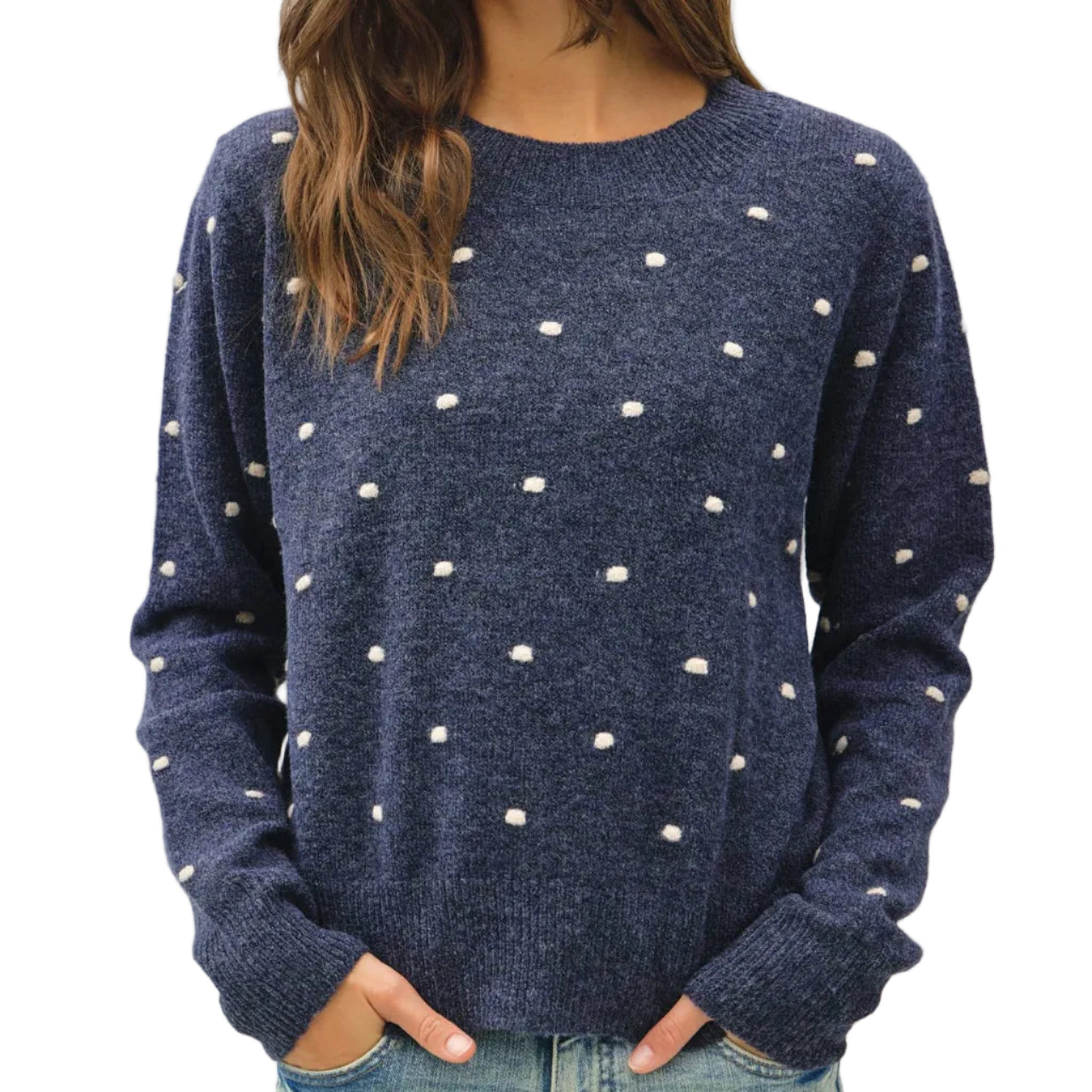 A woman wearing a navy blue knit sweater with small white polka dots, paired with light blue jeans, photographed against a plain light gray studio background.