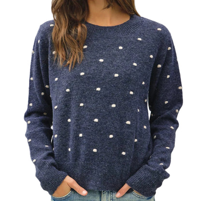 A woman wearing a navy blue knit sweater with small white polka dots, paired with light blue jeans, photographed against a plain light gray studio background.