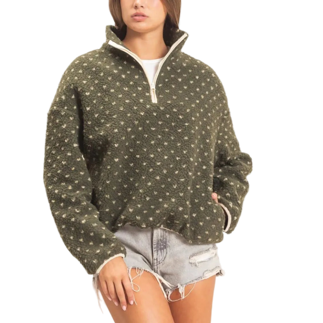 A woman modeling a dark olive green quarter-zip pullover sweater with a small white geometric pattern, paired with denim shorts and beige ankle boots, against a plain off-white studio background.