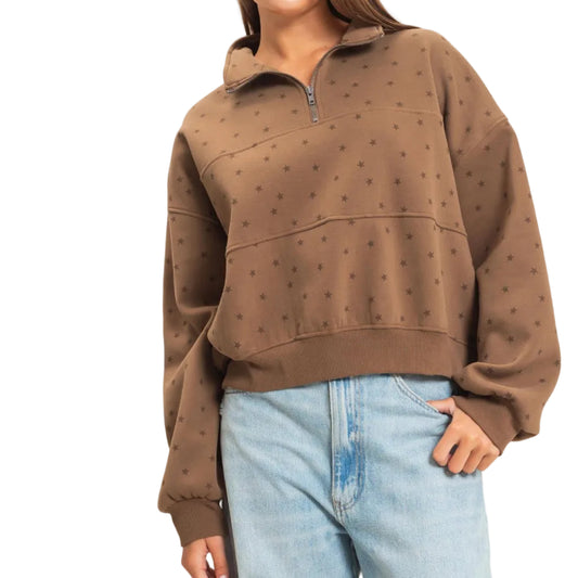 A woman wearing a brown cropped quarter-zip sweatshirt with a subtle polka dot pattern, paired with light-wash blue jeans, against a plain off-white background.