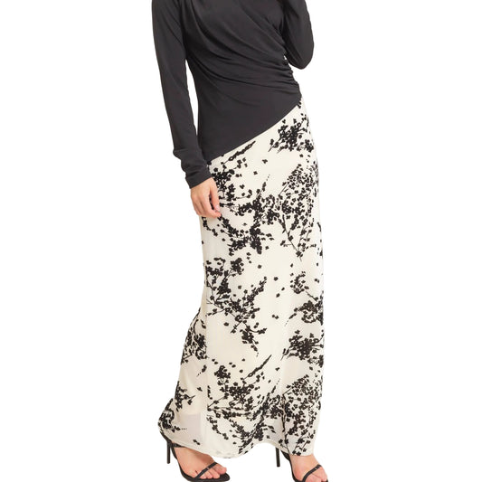 A model wearing a black long-sleeve top and a white maxi skirt with a black abstract floral print, paired with black high-heeled sandals, shown against a plain light background.