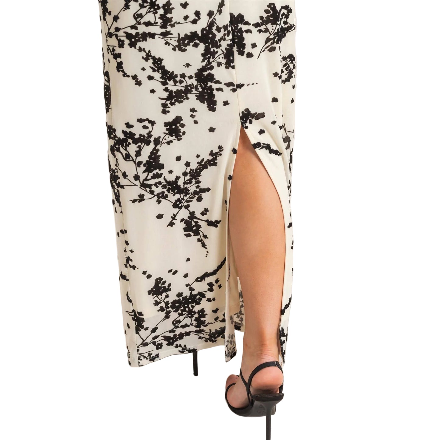 A close-up of a person wearing a long white maxi skirt with a black floral branch print and a high side slit, paired with black strappy high-heel sandals against a plain light background.
