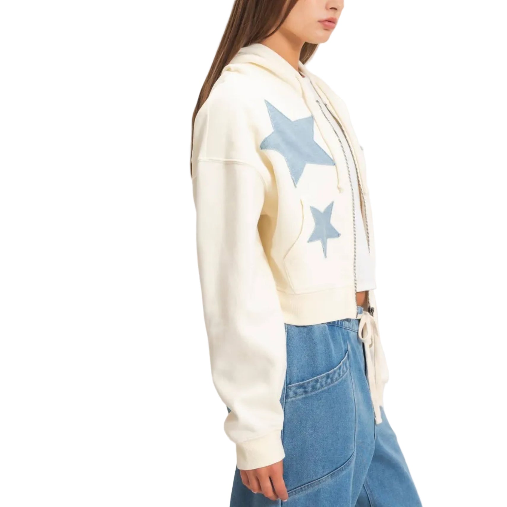 A woman wearing a cream-colored cropped hoodie with blue star patches on the front, paired with blue jeans, shown in a side profile against a light beige background.